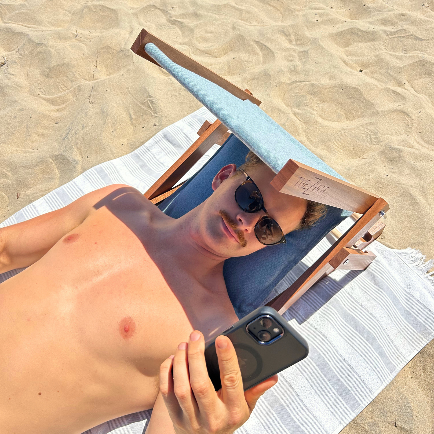 Person lying on a towel at the beach, using a phone with a mirror case.