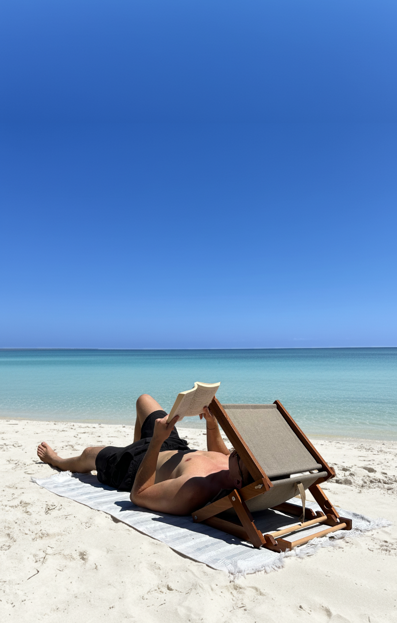 Person lying on a beach chair reading a book with clear blue sky and ocean in the background